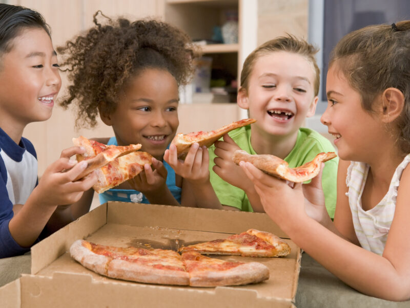 Four young children indoors eating pizza smiling